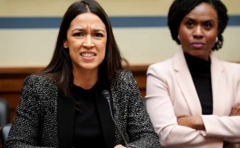 Alexandria Ocasio-Cortez speaking at a microphone with Ayanna Pressley seated behind her