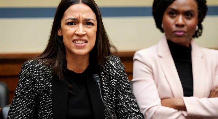 Alexandria Ocasio-Cortez speaking at a microphone with Ayanna Pressley seated behind her