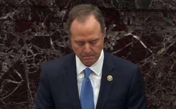 A man in a dark suit with a blue tie and congressional pin stands with his head bowed