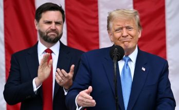Donald Trump speaking at a podium with JD Vance clapping behind him, American flag