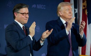 Two men in dark suits clapping on a stage with NRCC logo and American flag in background