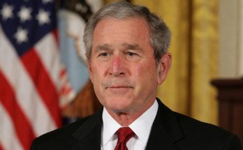 man in suit with American flag background, solemn expression, close-up