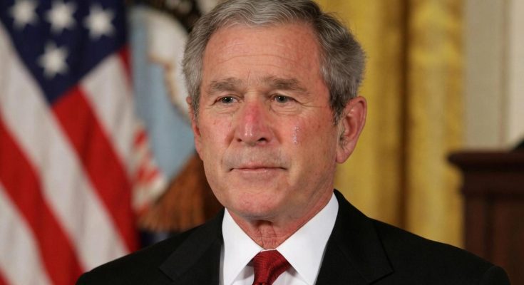 man in suit with American flag background, solemn expression, close-up