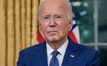 president Joe Biden in blue suit and tie seated at desk with gold curtains and American