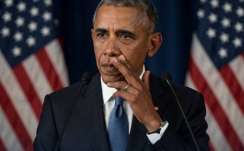 barack Obama speaking at podium with American flags behind, touching face with concerned