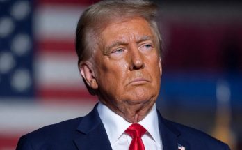 donald Trump in navy suit with red tie stands before American flag with Emergency News