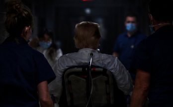 patient in wheelchair escorted by medical staff in dim hallway