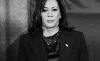 woman in dark suit with flag pin and necklace, solemn expression