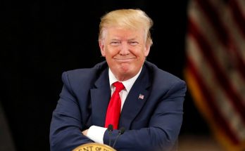 donald Trump smiles with arms crossed at presidential podium with American flag behind