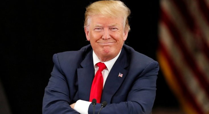 donald Trump smiles with arms crossed at presidential podium with American flag behind