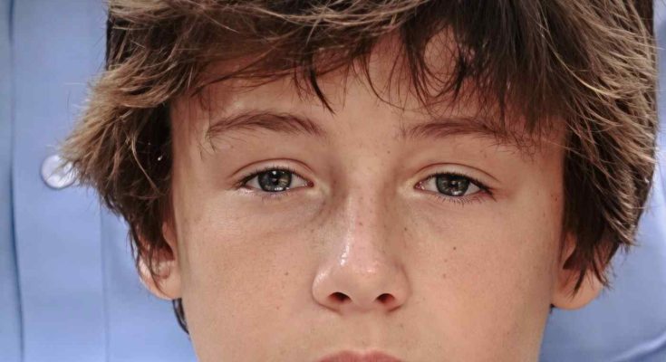 close-up portrait of a teenage boy with tousled brown hair and freckles, wearing a