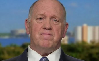 middle-aged man in navy suit and striped tie against blurred waterfront cityscape with