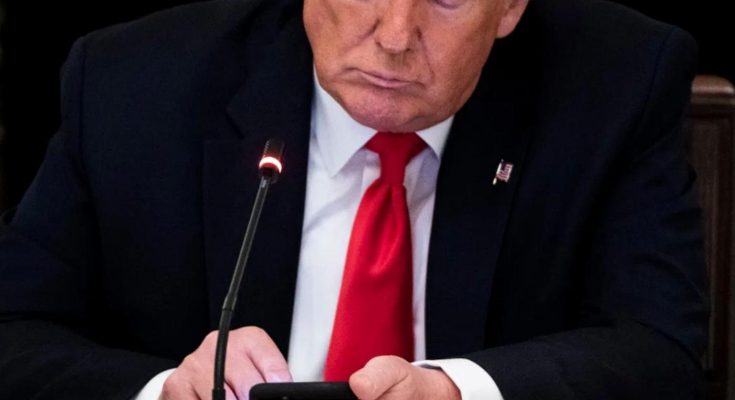 donald Trump in dark suit and red tie looking down at smartphone while seated at desk