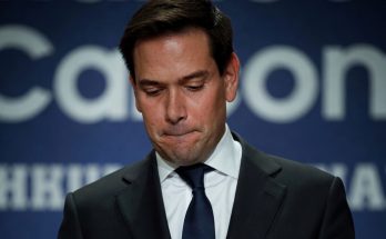 marco Rubio looking down solemnly at podium with Breaking News banner overlay