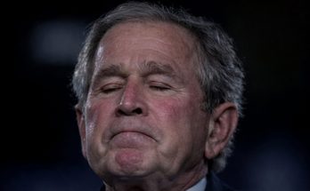 george W. Bush with eyes closed and somber expression against dark background with yellow