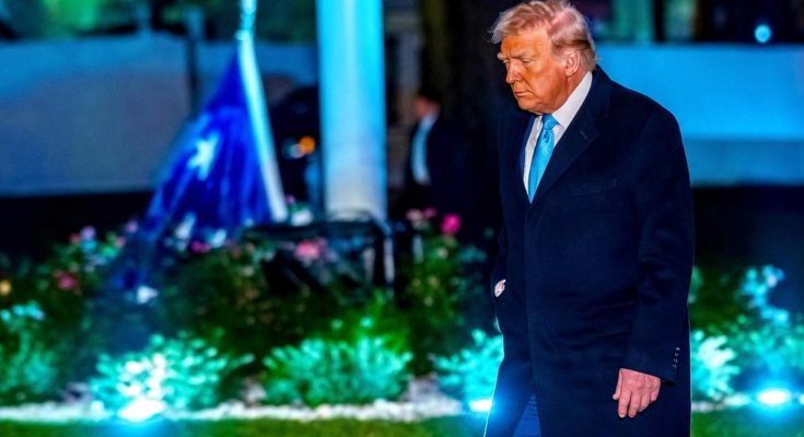 donald Trump walks across a lawn at night wearing a dark overcoat and light blue tie