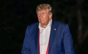 donald Trump in navy suit with loosened red tie holding red MAGA hat at night