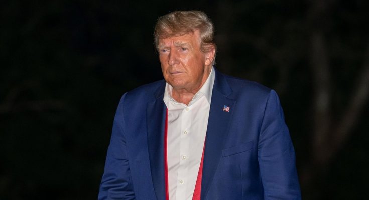 donald Trump in navy suit with loosened red tie holding red MAGA hat at night
