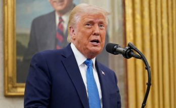 donald Trump speaking at podium with blue tie, portrait of George W. Bush visible behind
