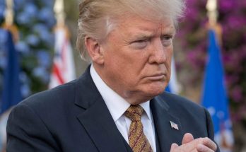 donald Trump in dark suit with patterned gold tie, hands clasped, with overlaid text