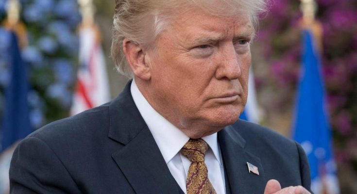 donald Trump in dark suit with patterned gold tie, hands clasped, with overlaid text