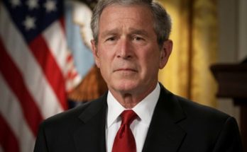 former President George W. Bush in dark suit and red tie stands before American flag with