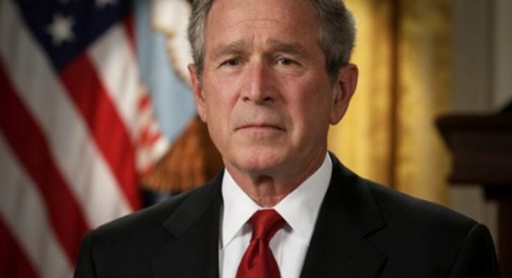 former President George W. Bush in dark suit and red tie stands before American flag with