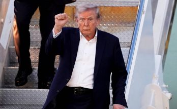 donald Trump raises clenched fist while descending airplane stairs in navy suit