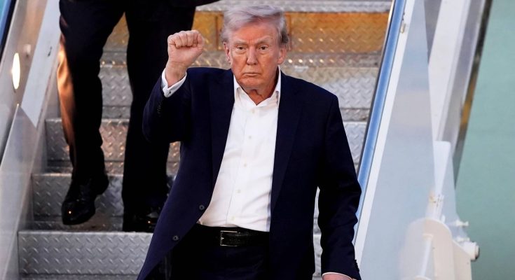 donald Trump raises clenched fist while descending airplane stairs in navy suit