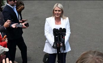 blonde woman in white suit addresses press outside with multiple microphones