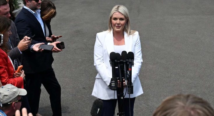 blonde woman in white suit addresses press outside with multiple microphones