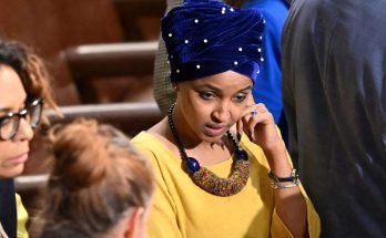 ilhan Omar wearing yellow top and blue velvet turban with pearl studs, seated in