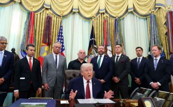 donald Trump seated at Oval Office desk gesturing while speaking, surrounded by officials