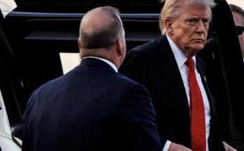 donald Trump wearing dark suit and red tie exits vehicle with Secret Service agent nearby
