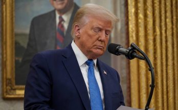 donald Trump reading from notes at a podium with breaking news banner and portrait of