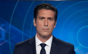news anchor in dark suit with silver tie clip delivering breaking news in television