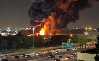 massive industrial fire erupts alongside busy highway at night, sending towering black