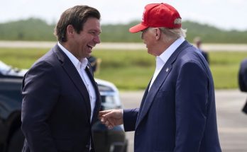 donald Trump in red MAGA cap shakes hands with Ron DeSantis on airport tarmac with black