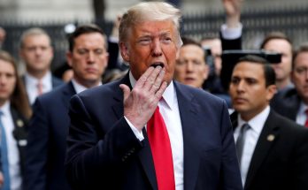 donald Trump in navy suit and red tie speaking outdoors with hand near mouth, surrounded