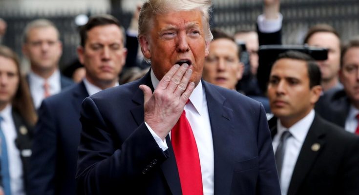 donald Trump in navy suit and red tie speaking outdoors with hand near mouth, surrounded