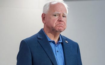 tim Walz stands in a blue blazer and polo shirt with a somber expression against a plain