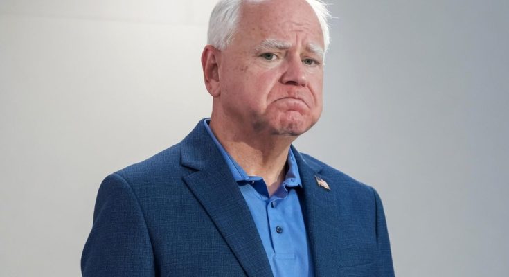 tim Walz stands in a blue blazer and polo shirt with a somber expression against a plain