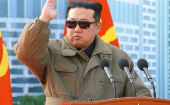 kim Jong Un salutes at wooden podium with hammer and sickle emblem, wearing olive jacket