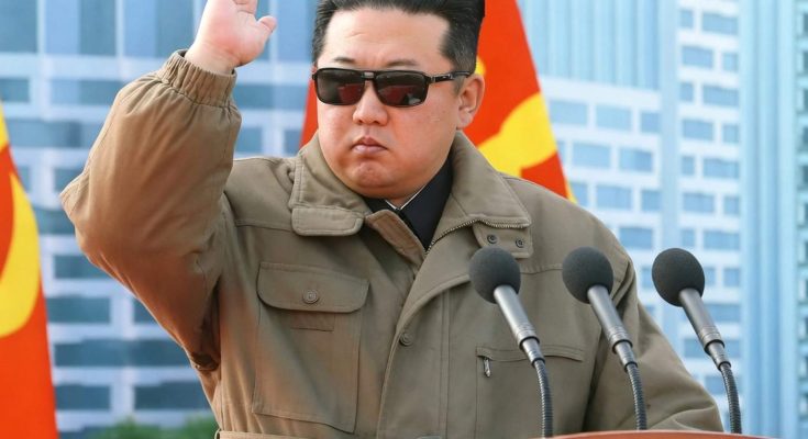 kim Jong Un salutes at wooden podium with hammer and sickle emblem, wearing olive jacket